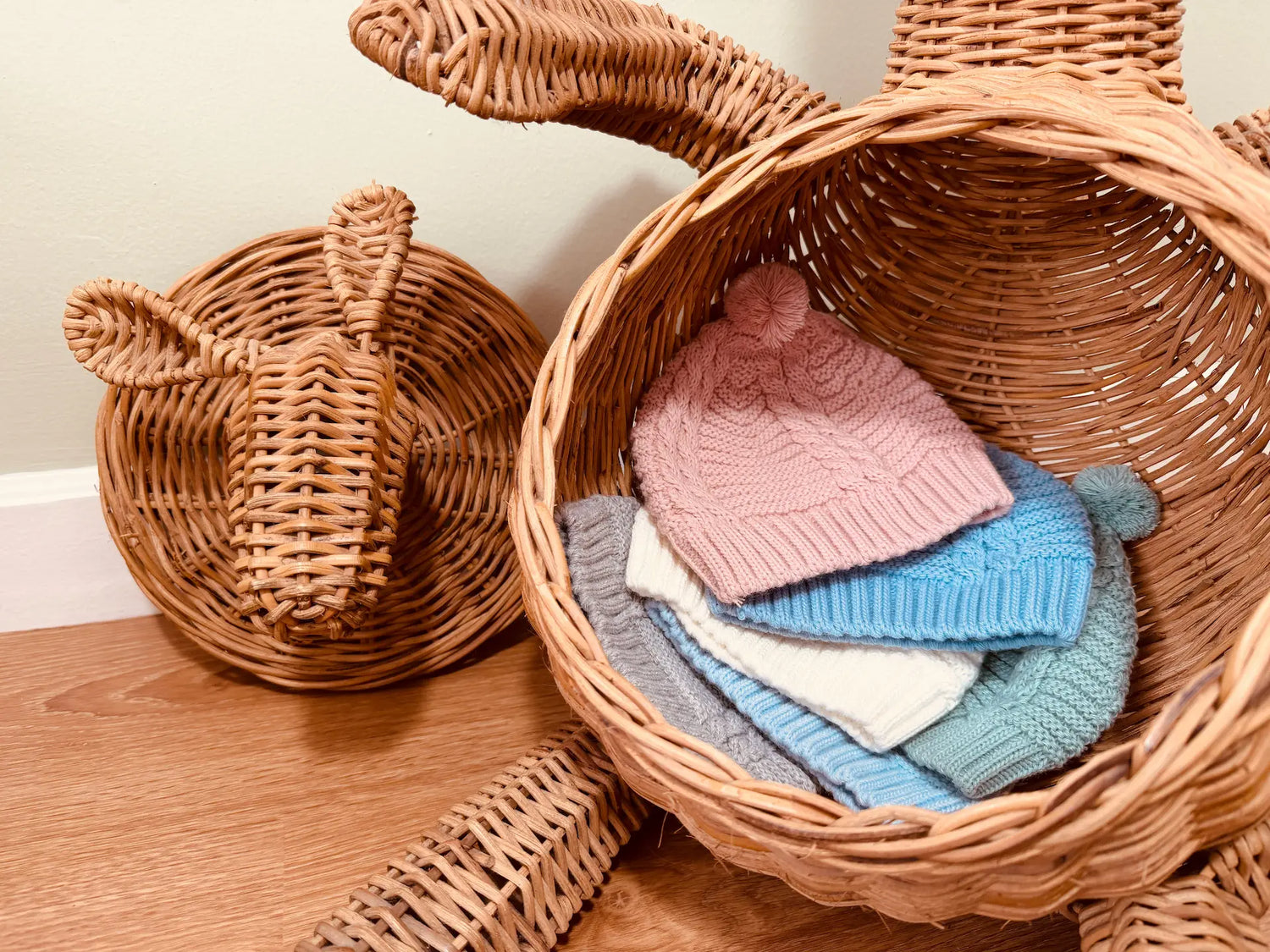 Seis variantes del gorro de punto trenzado con pompón en los colores rosa, cobalto, perla, verde, azul y gris dentro de la barriga de un conejo grande de mimbre