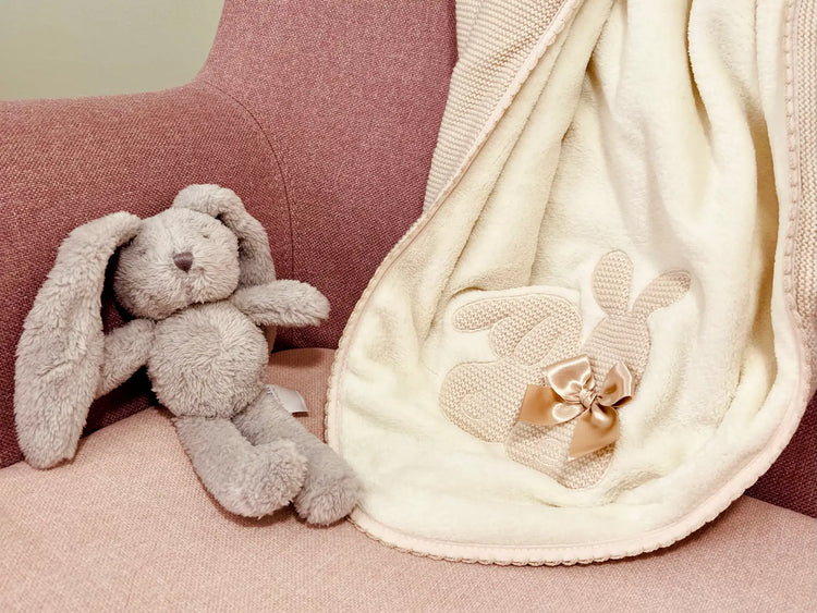 Manta con motivo de conejos de punto y lazo en color beige dispuesta sobre un sillón con un conejito de peluche al lado