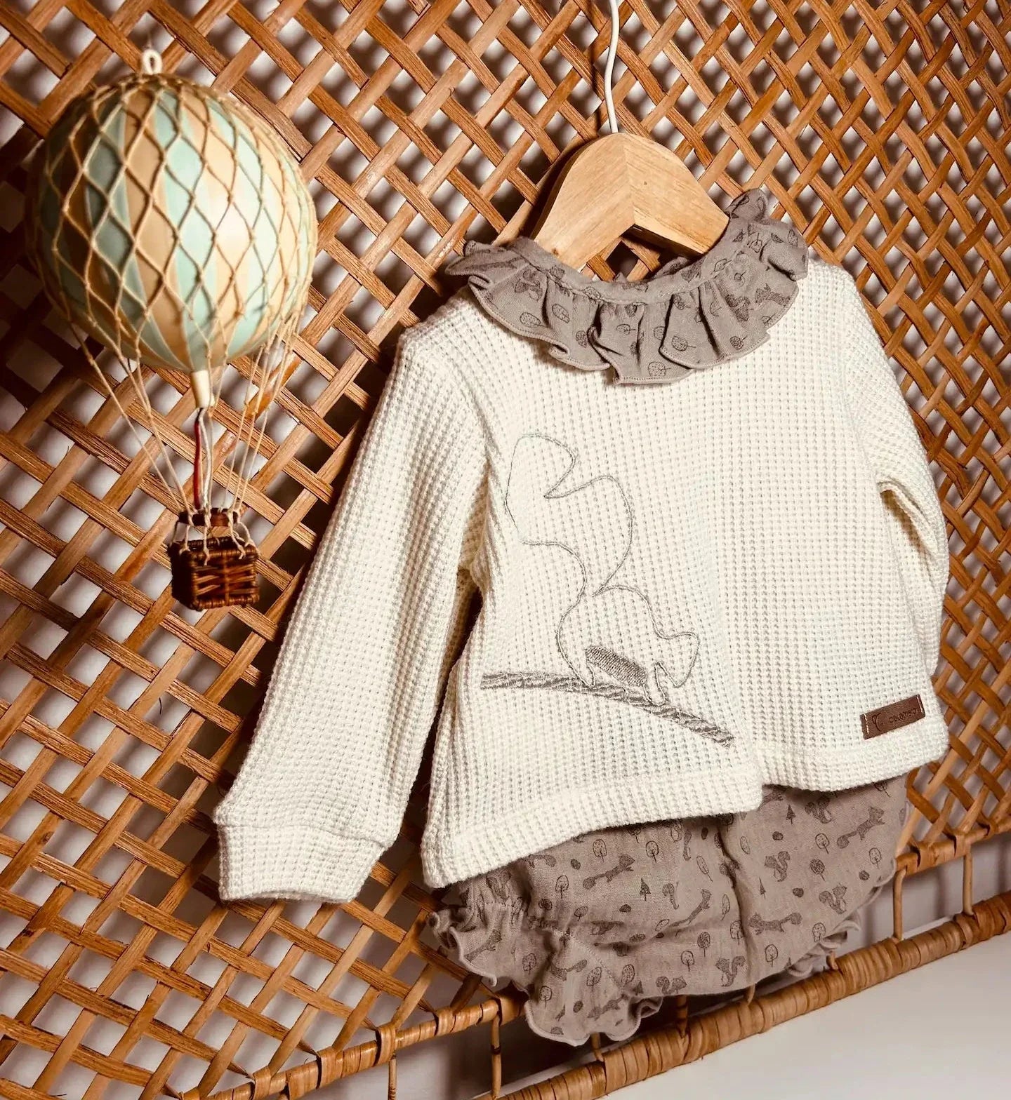 Conjunto de niña de camiseta con volante en el cuello y cubrepañal colgado en un soporte de mimbre con un globo aerostático de juguete al lado