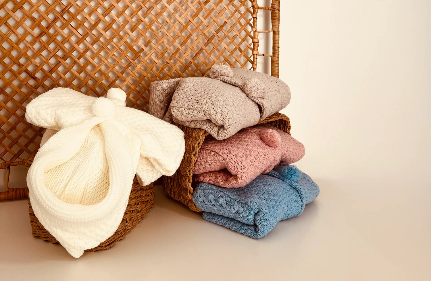 Cuatro variantes de chaqueta de punto con capucha y pompones en la zona de los botones en perla, topo, rosa y cobalto de la colección de lanzamiento de Lah Lah en cestas de mimbre